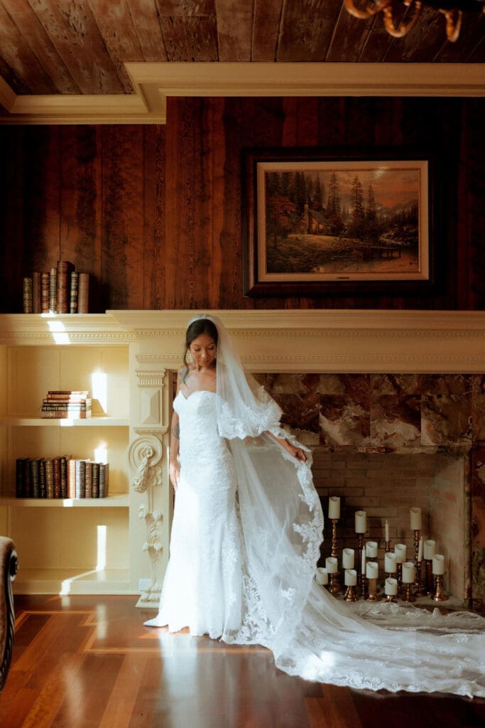 Bride stands beside a grand fireplace while adjusting her lace veil before her wedding ceremony in Mobile, Alabama, photographed in a timeless, film-inspired style.