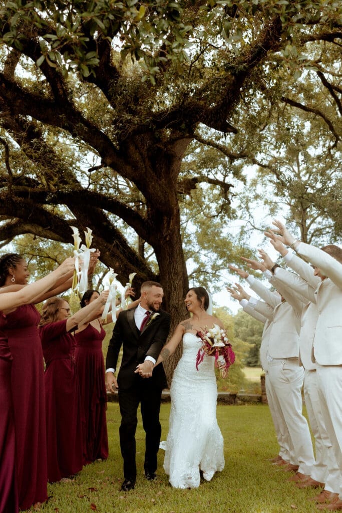 Newlyweds walk hand in hand through a ribbon exit surrounded by family and friends after an outdoor wedding ceremony in Mobile, Alabama, photographed in a documentary, film-inspired style.