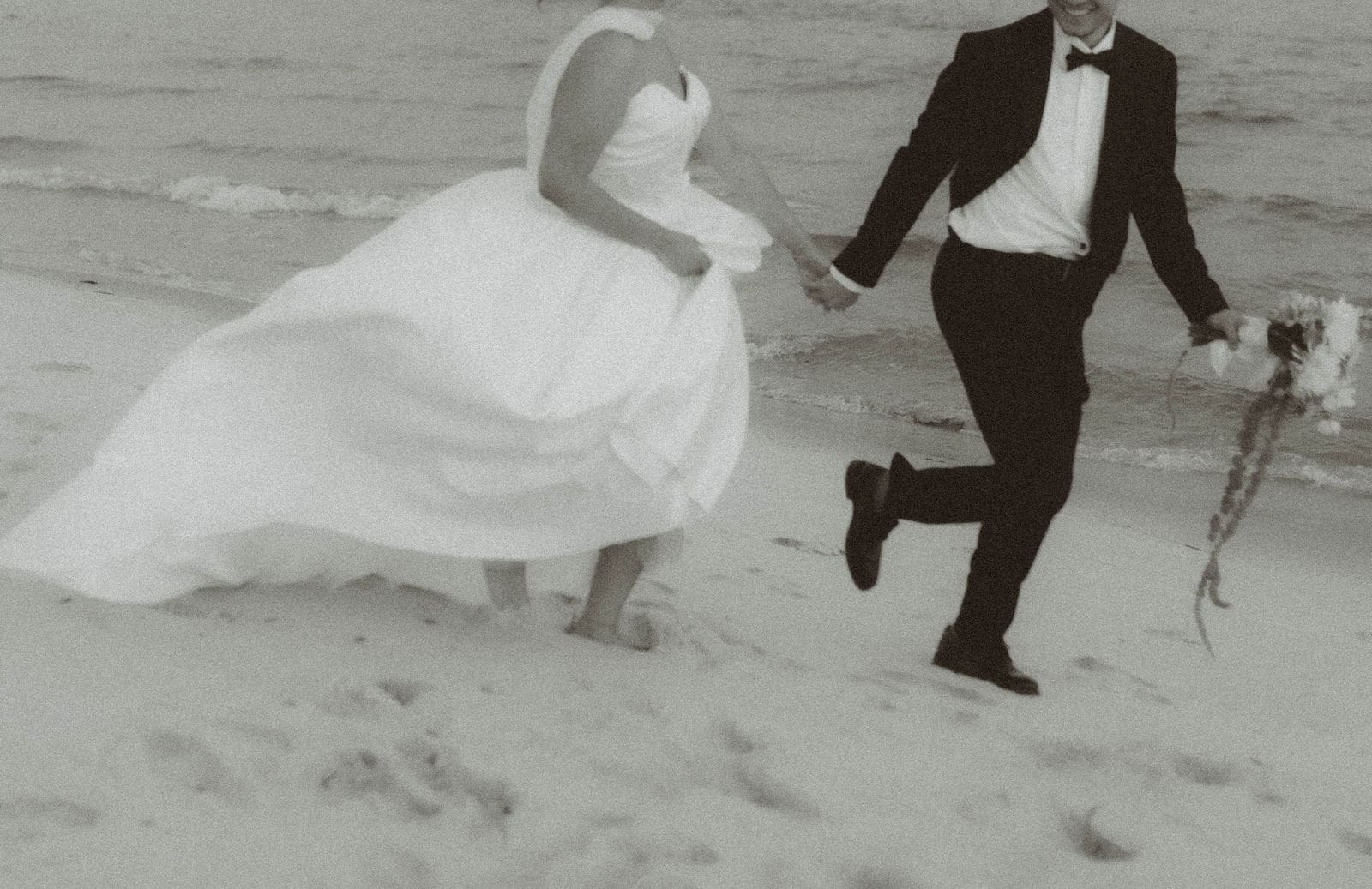 Black and white photo of a newlywed couple running along the beach during their Gulf Coast wedding
